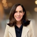 Headshot of a person with shoulder-length dark hair, smiling, wearing a beige jacket over a black shirt, with a blurred background.