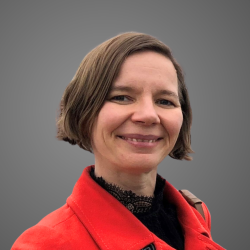 A person smiling, wearing a red coat and a black lace top, with short brown hair against a neutral gray background.