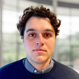 A headshot of a person with curly hair, wearing a navy sweater over a checkered shirt. The background is out of focus.