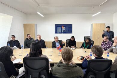 A small group of people sitting around a conference table in a meeting room. One person appears to be speaking, while others listen or take notes. There is a sign on the wall indicating this may be a formal discussion setting.