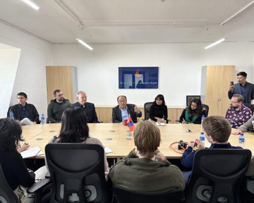 A small group of people sitting around a conference table in a meeting room. One person appears to be speaking, while others listen or take notes. There is a sign on the wall indicating this may be a formal discussion setting.