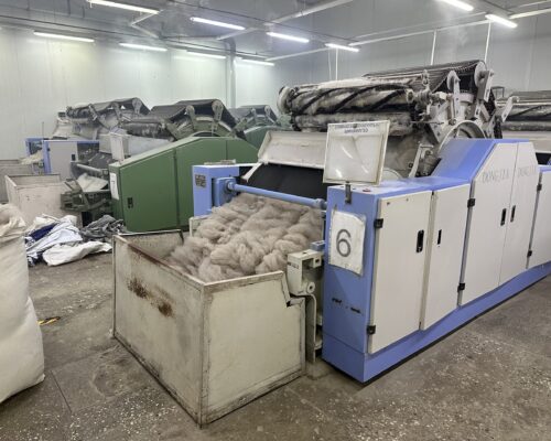 The image shows industrial machinery in a textile factory, with wool or fiber being processed in large machines. The environment appears industrial with several machines and large containers of fibers.