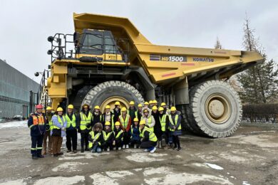 A large group of people wearing safety vests and helmets stand in front of a massive yellow Komatsu HD1500 dump truck. The setting appears to be an industrial or construction site.