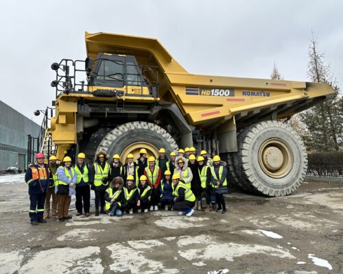 A large group of people wearing safety vests and helmets stand in front of a massive yellow Komatsu HD1500 dump truck. The setting appears to be an industrial or construction site.