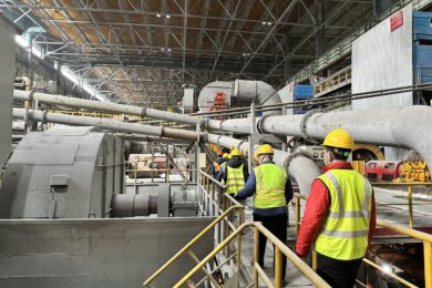 A group of people wearing yellow hard hats and reflective vests walk through an industrial facility with large machinery and pipes. The setting is a spacious factory or plant.