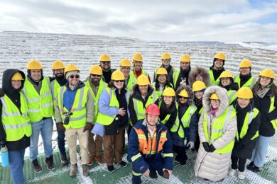 A group of people wearing yellow hard hats and high-visibility vests stand outdoors on a snowy day, possibly visiting a construction or mining site.