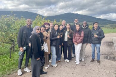 A group of people standing outdoors in a vineyard, smiling and holding wine glasses. They are dressed in casual, warm clothing with hills in the background under a cloudy sky.