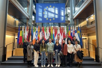 A group of people stand in front of a display of various national flags and a large European Union flag in a spacious, modern building with high ceilings and glass features.