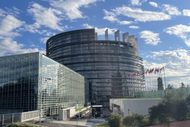 A modern building with circular and glass architecture against a blue sky with clouds. Flags are displayed along the right side of the structure.