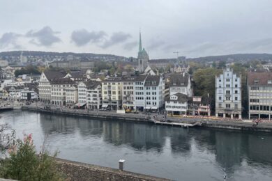 A cityscape view with a river in the foreground, lined with historic buildings featuring varied architectural styles. A church with a green spire is prominent in the background under a cloudy sky.