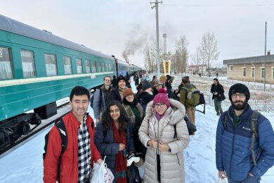 A small group of people standing on a snow-covered train platform, smiling and dressed in winter clothing. A green train is visible on the left.