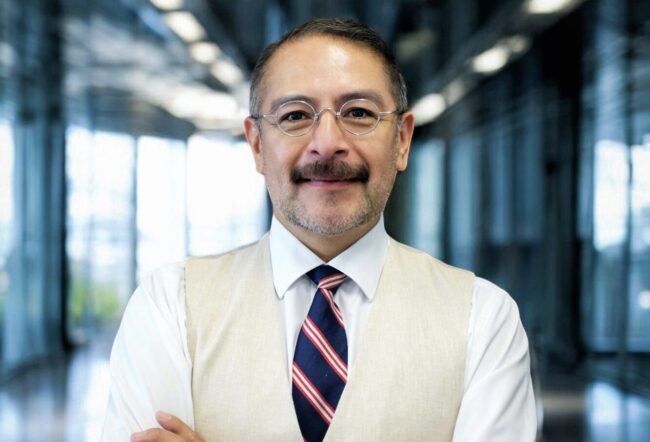 A person wearing glasses, a white shirt, a beige vest, and a striped tie stands confidently with arms crossed in a modern, glass-paneled hallway.