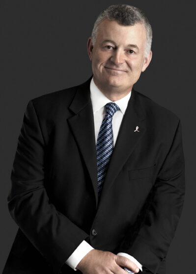 A headshot of a person wearing a dark suit, white shirt, and patterned tie, with a subtle pink ribbon pin on the lapel, against a dark background.