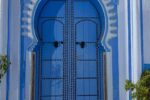 An ornate blue door with intricate patterns set in an archway, framed by white and blue walls. A hanging lantern is above the door.