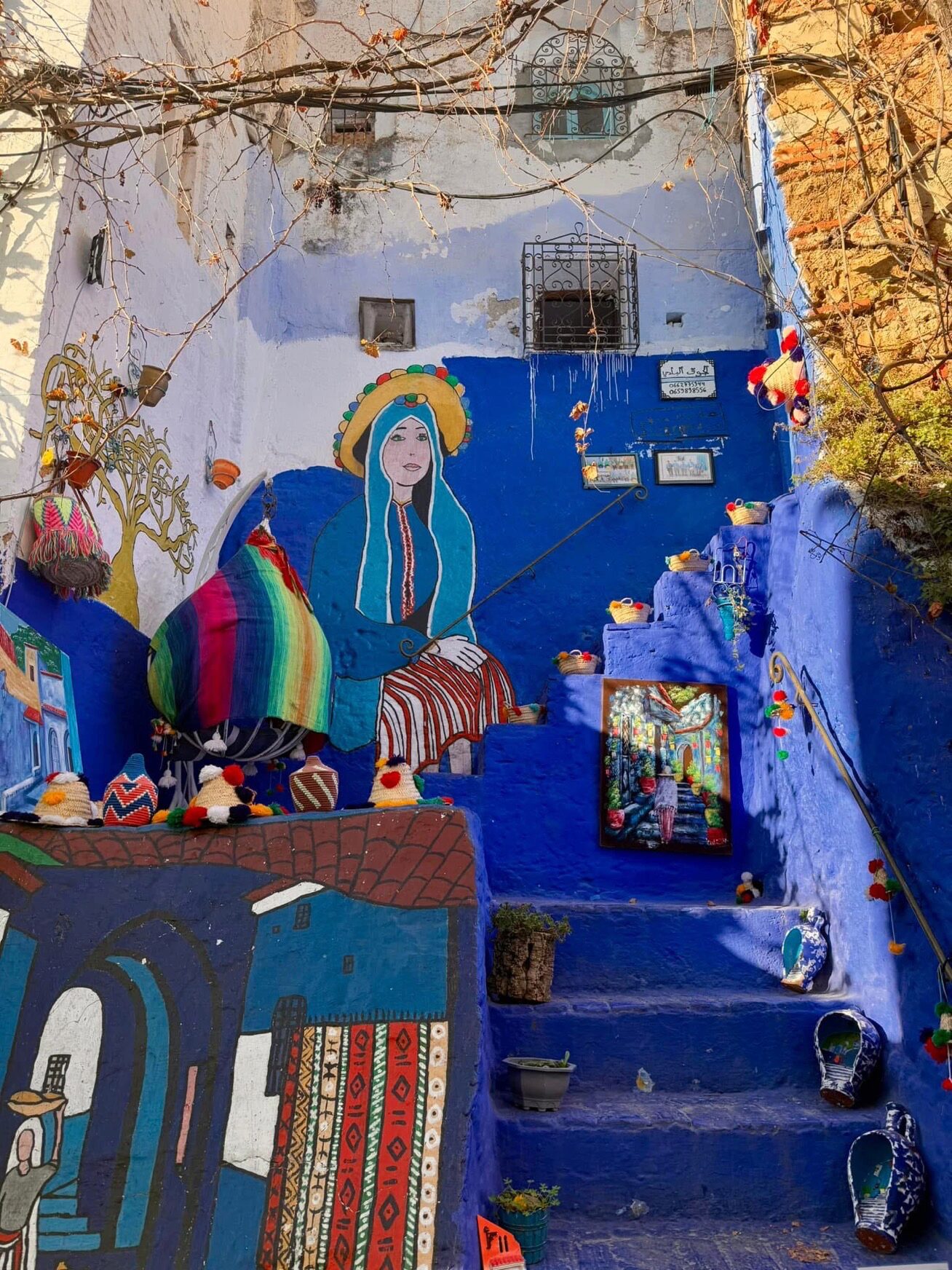 Colorful street art on blue walls, featuring a large mural of a person in traditional attire, vibrant tapestries, and painted pots on stairs.