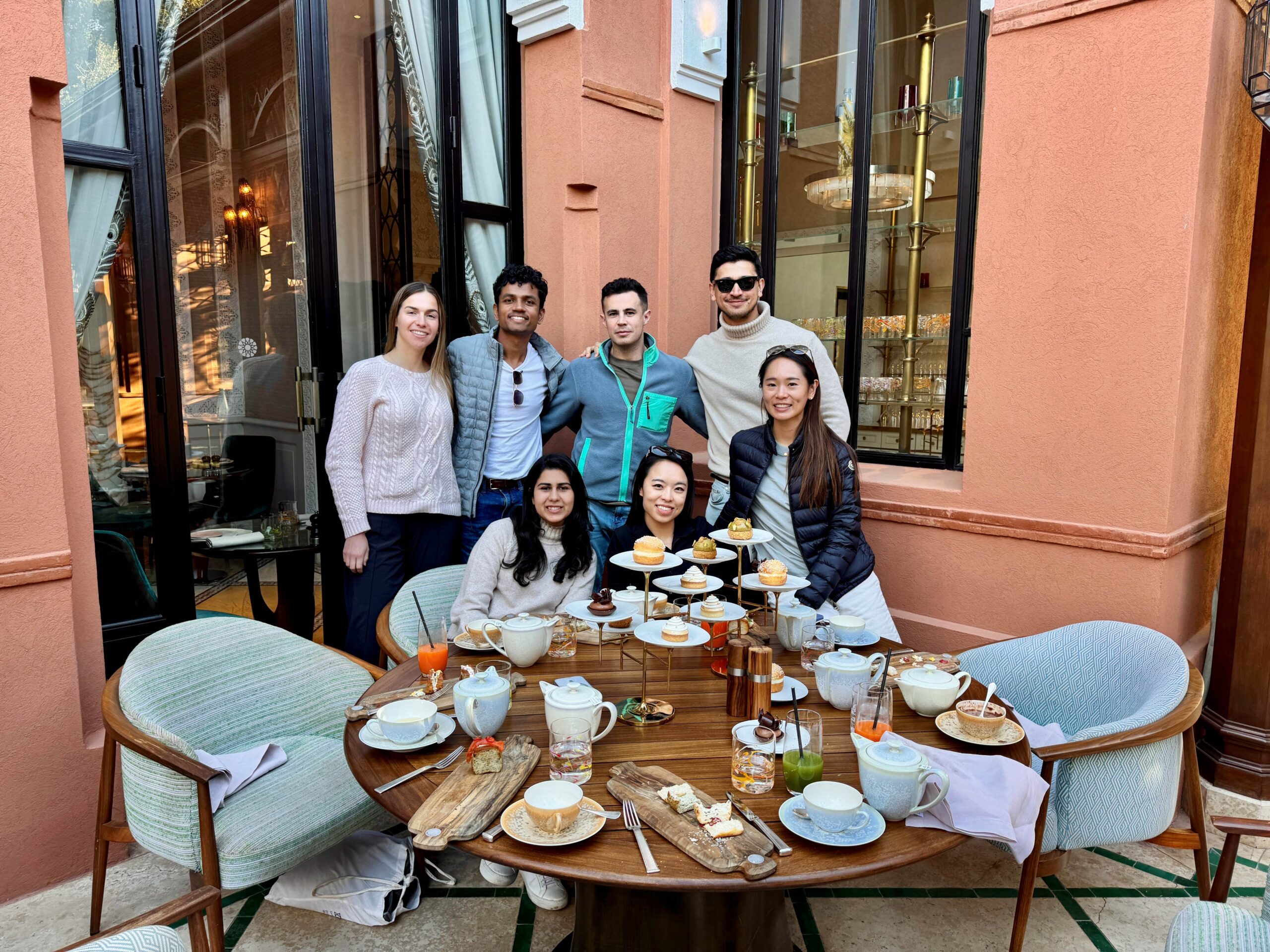 A group of people posing for a photo around a table set with tea, pastries, and appetizers. They are outside a building with large windows.