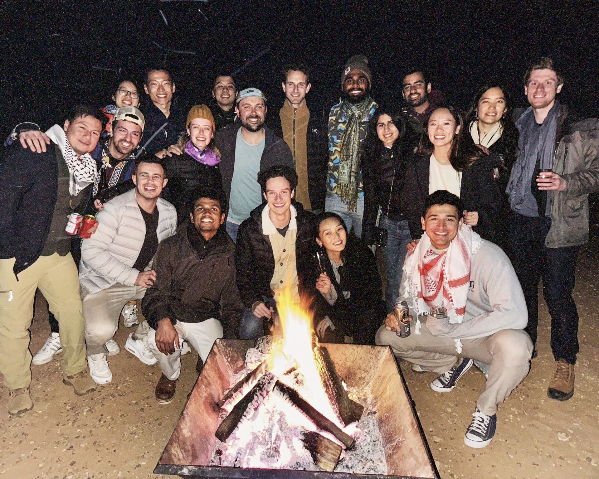 A group of people gathered around a campfire at night, smiling and enjoying a casual outdoor setting. Some are holding drinks and dressed in warm clothing.