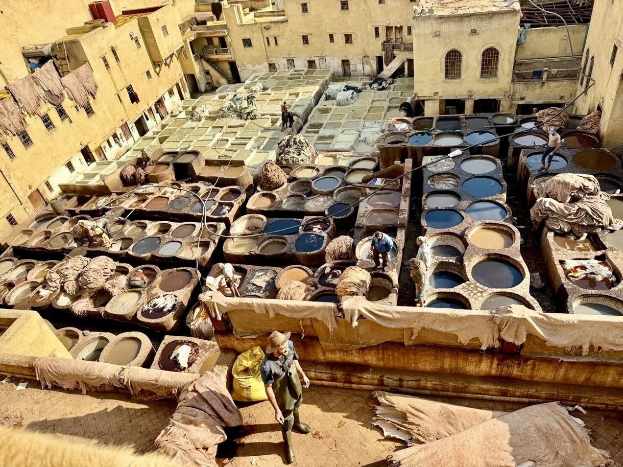 A traditional tannery with workers processing leather in a series of stone vats filled with various colored liquids and hides, surrounded by yellow buildings.