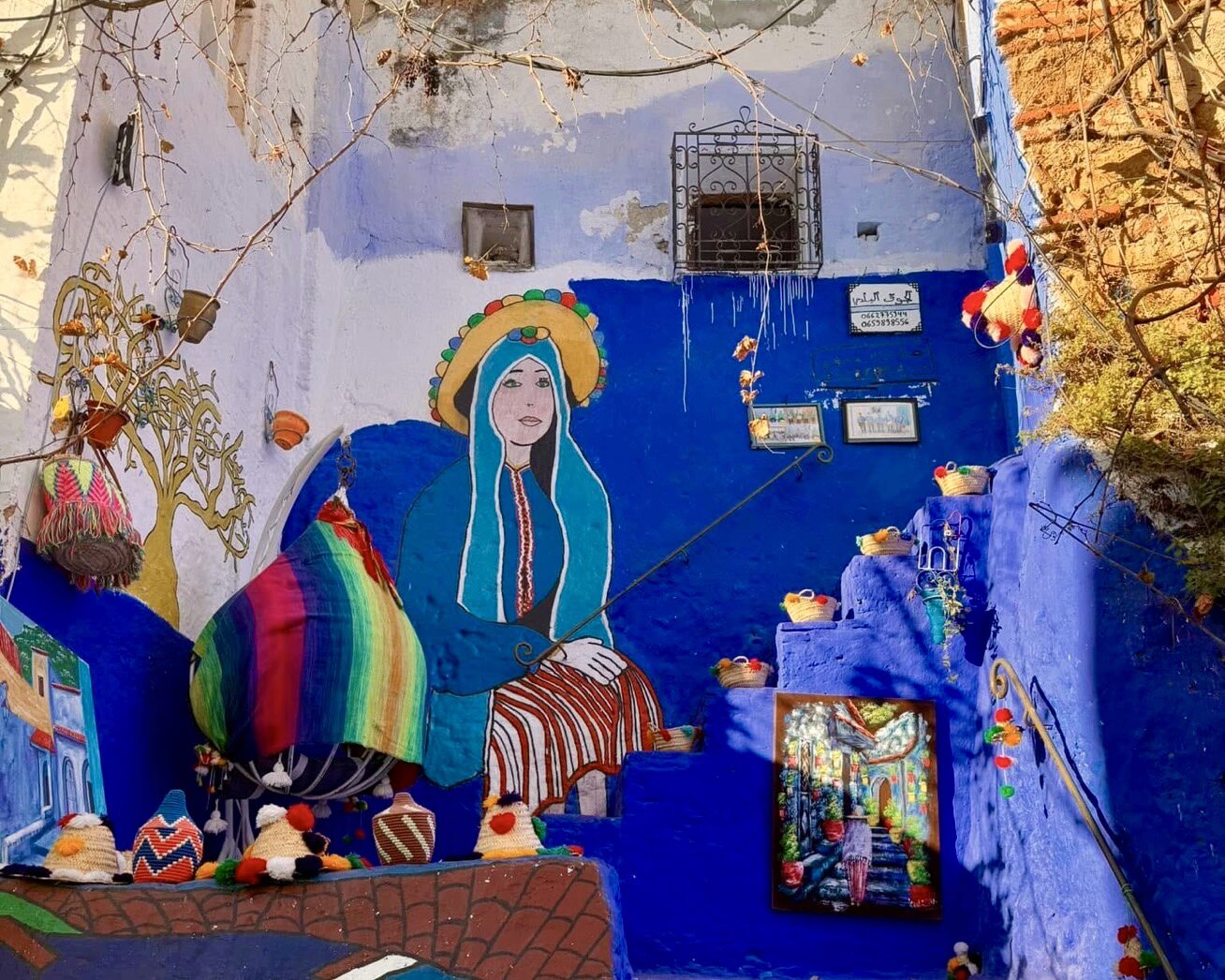 A vibrant street art mural featuring a person in traditional clothing painted on a blue staircase. The scene includes colorful woven baskets, fabric, and framed art, blending cultural and artistic elements.