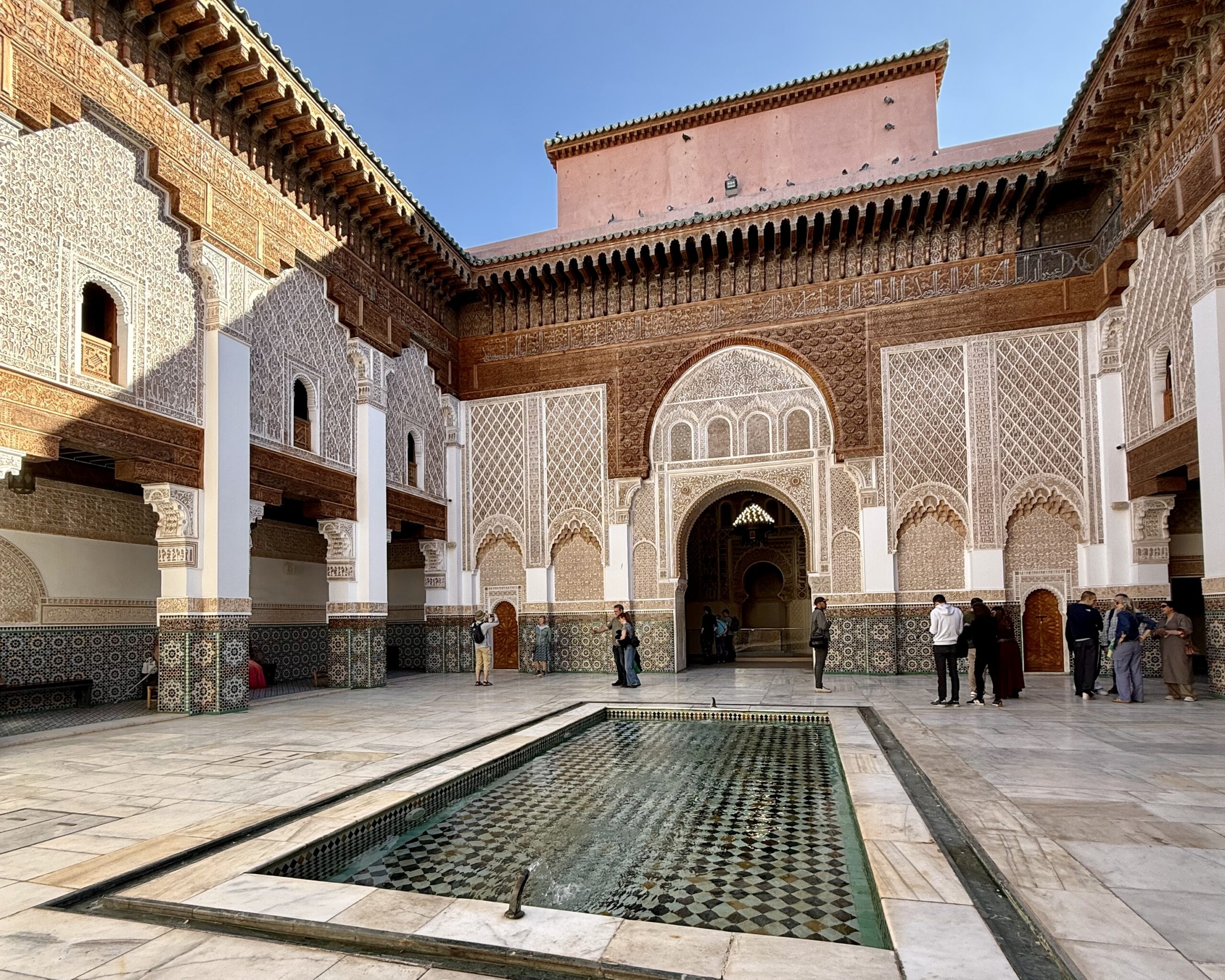 The image depicts a courtyard of a traditional Islamic architecture style, featuring intricately carved arches, patterned tile work, and a rectangular reflecting pool. Several people are walking around and admiring the details.