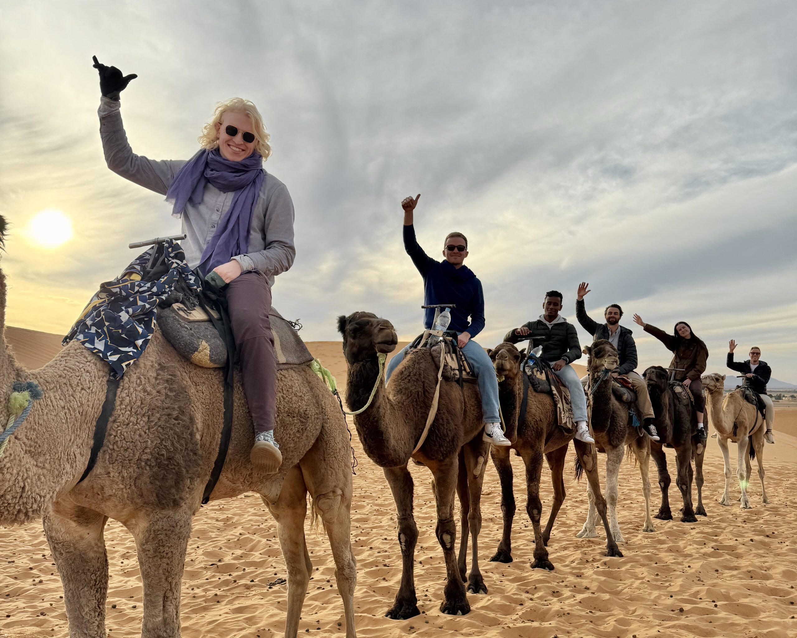 People riding camels in a desert setting, with a bright sky and sand dunes in the background. They are smiling and waving, suggesting a friendly and adventurous atmosphere.