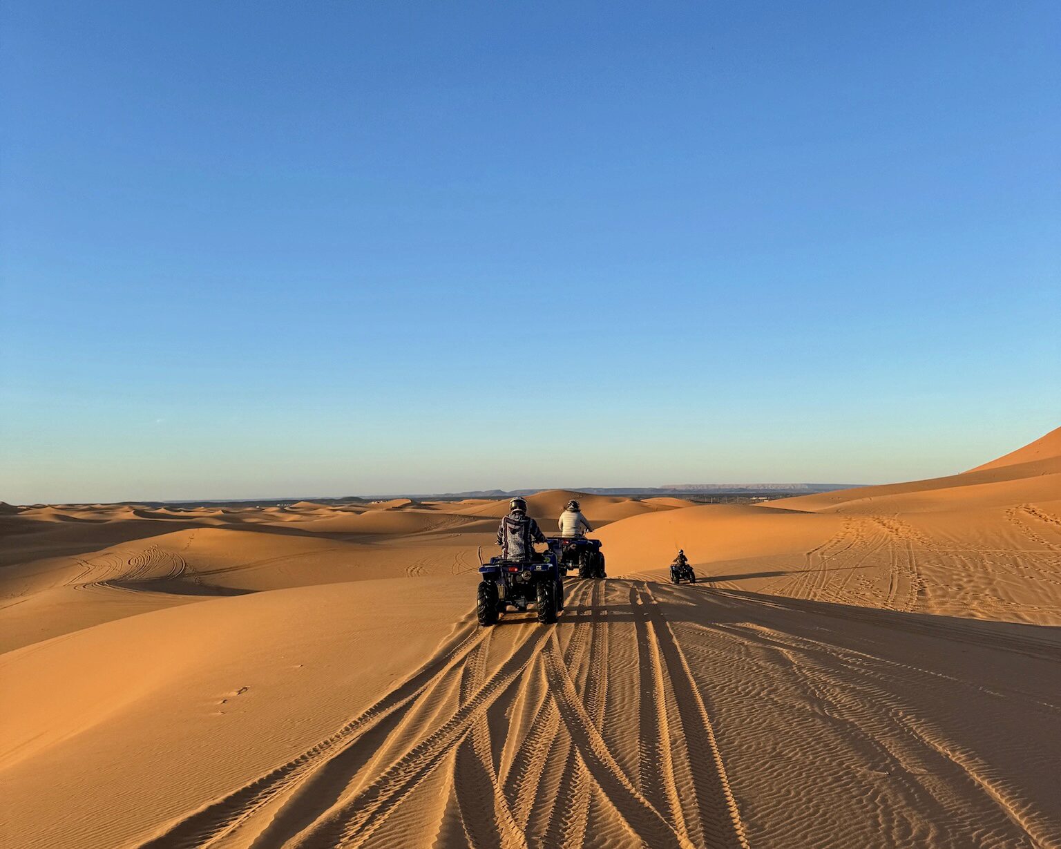 Riders on ATVs traverse sandy dunes under a clear blue sky, with tire tracks visible on the desert landscape.