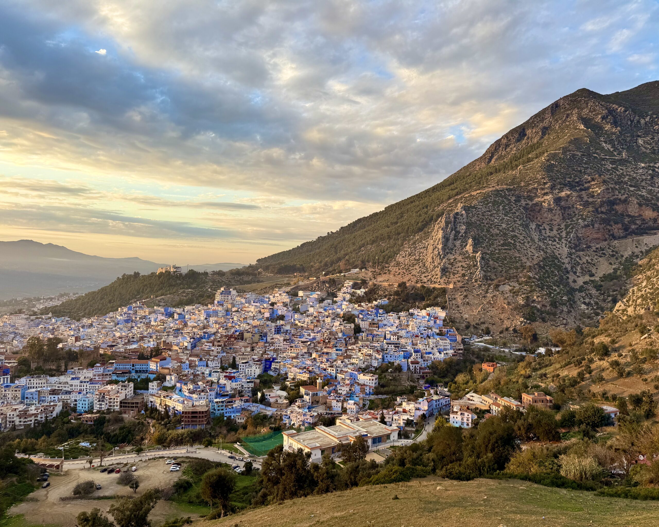 A scenic view of Chefchaouen, a town in Morocco known for its blue-washed buildings. It is set against a backdrop of rolling hills and a dramatic mountain under a partly cloudy sky.