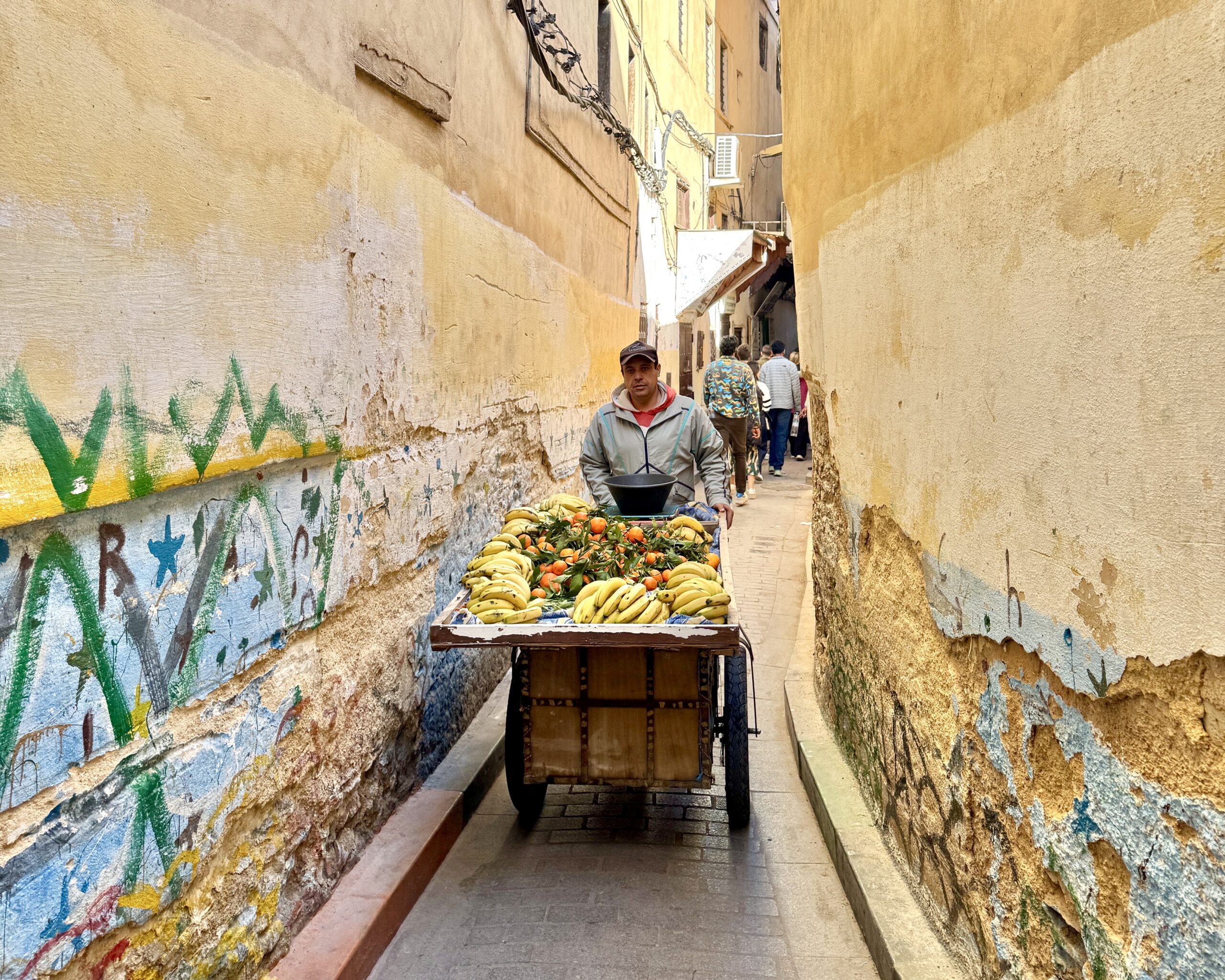 A vendor in a narrow alleyway pushing a fruit cart filled with bananas and oranges. The alley walls are weathered and partially covered in graffiti.