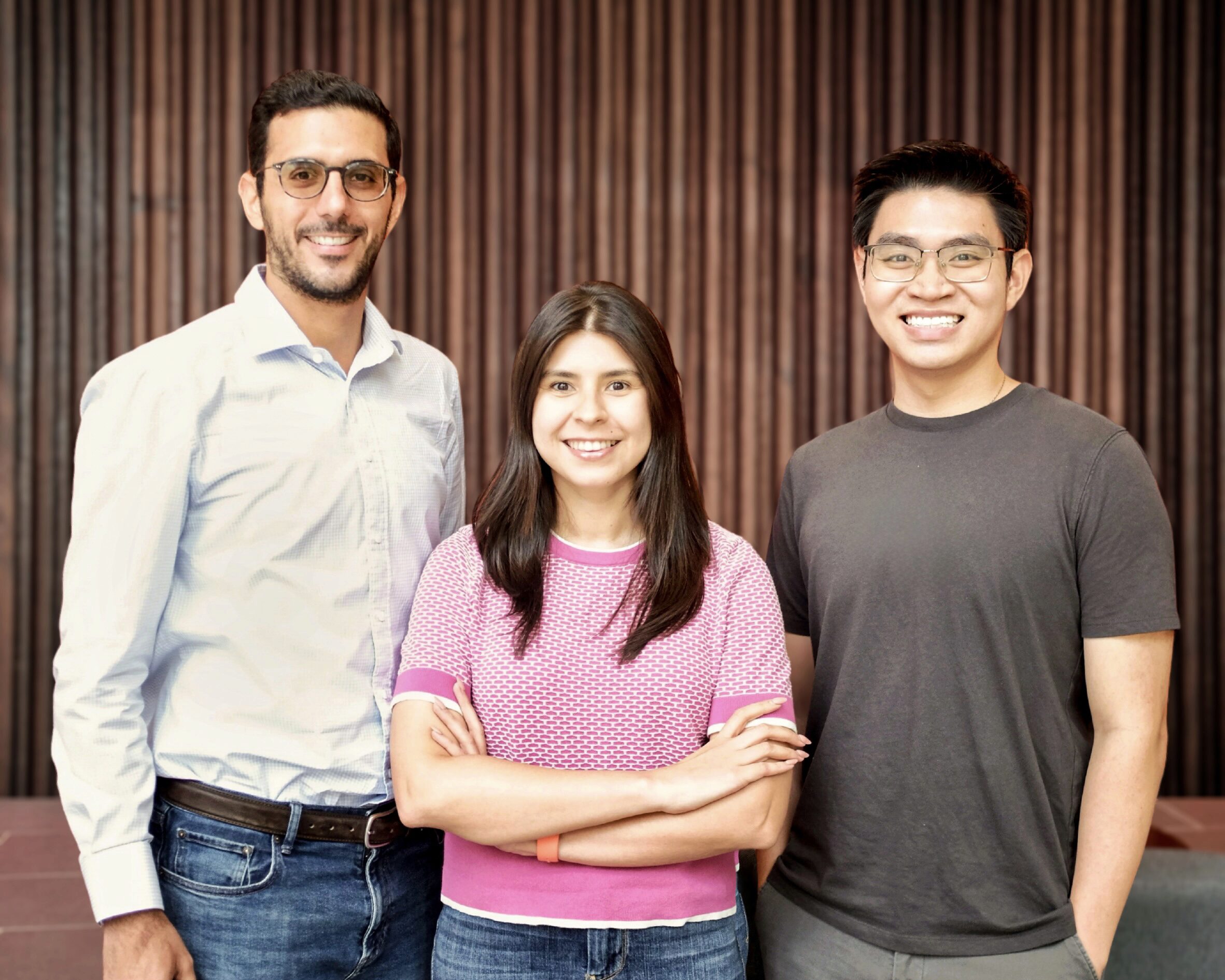 A small group of three people standing together, smiling, indoors against a striped wooden background. The person in the middle has their arms crossed.