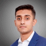 Headshot of a person wearing a blue suit and white shirt, with a neutral expression, against a gray background.