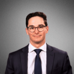 Headshot of a person wearing glasses and a suit, set against a plain gray background.