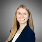 headshot of person in professional attire with long blonde hair, smiling against a gray background.