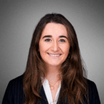 A person with long dark hair is smiling, wearing a striped suit jacket and a white shirt, in front of a gray background.