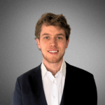 A person with short wavy hair is wearing a suit and white shirt, standing against a plain gray background.