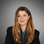 Headshot of a person with long hair, wearing a blazer against a plain gray background.
