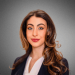 Dina Daas Headshot of a person with long, wavy hair wearing a blazer and a white top, smiling against a gray background.