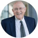 Headshot of a person wearing glasses, a suit, and a patterned tie, smiling in an indoor setting.