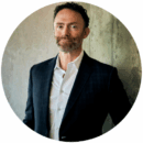 Headshot of person in a suit leaning against a concrete wall, with a confident expression.