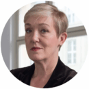 Headshot of a person with short, light-colored hair, wearing a dark jacket, in front of a window.