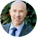 Headshot of a person smiling, wearing a suit and tie, with greenery in the background.