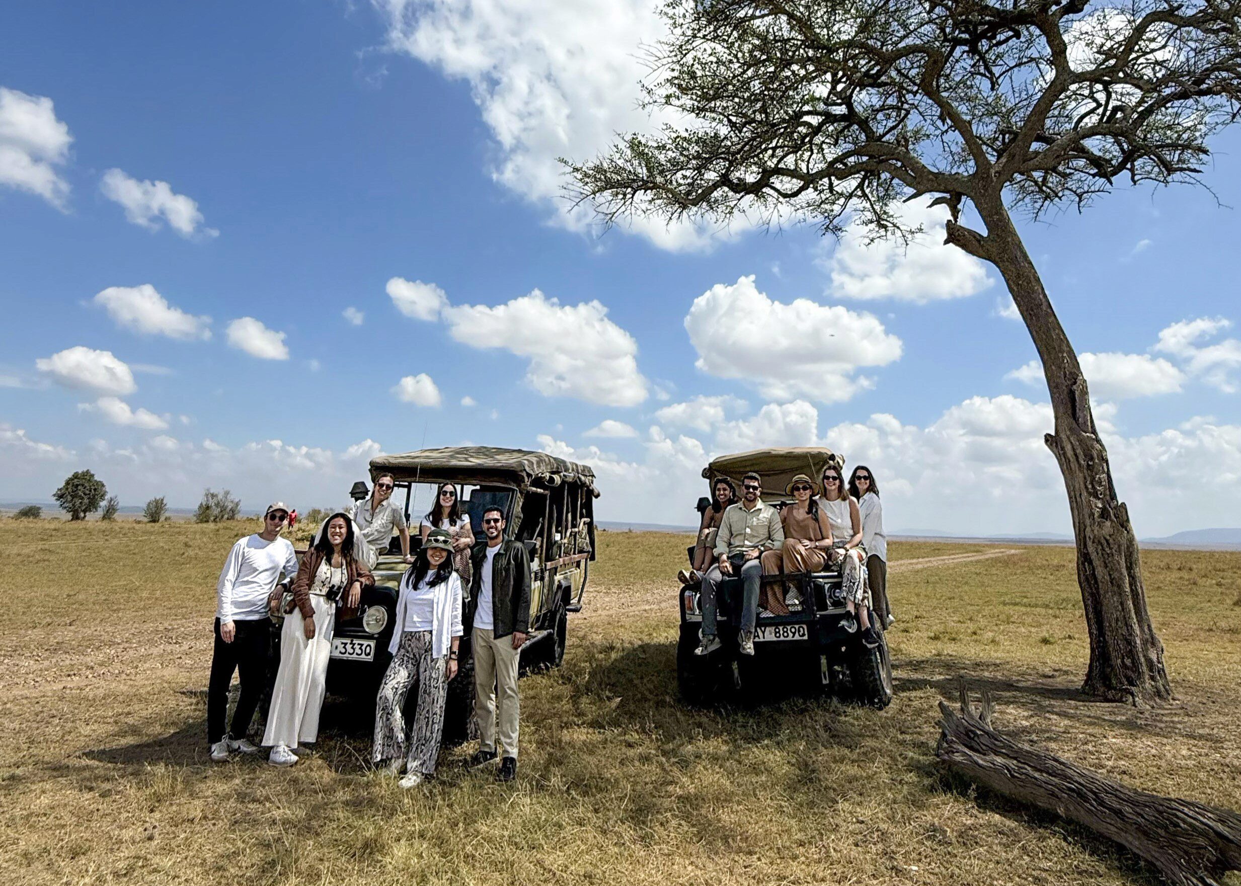 Culture Quest 2025 A group of people on a safari, standing and sitting beside two open-top vehicles in a savannah landscape with an acacia tree.
