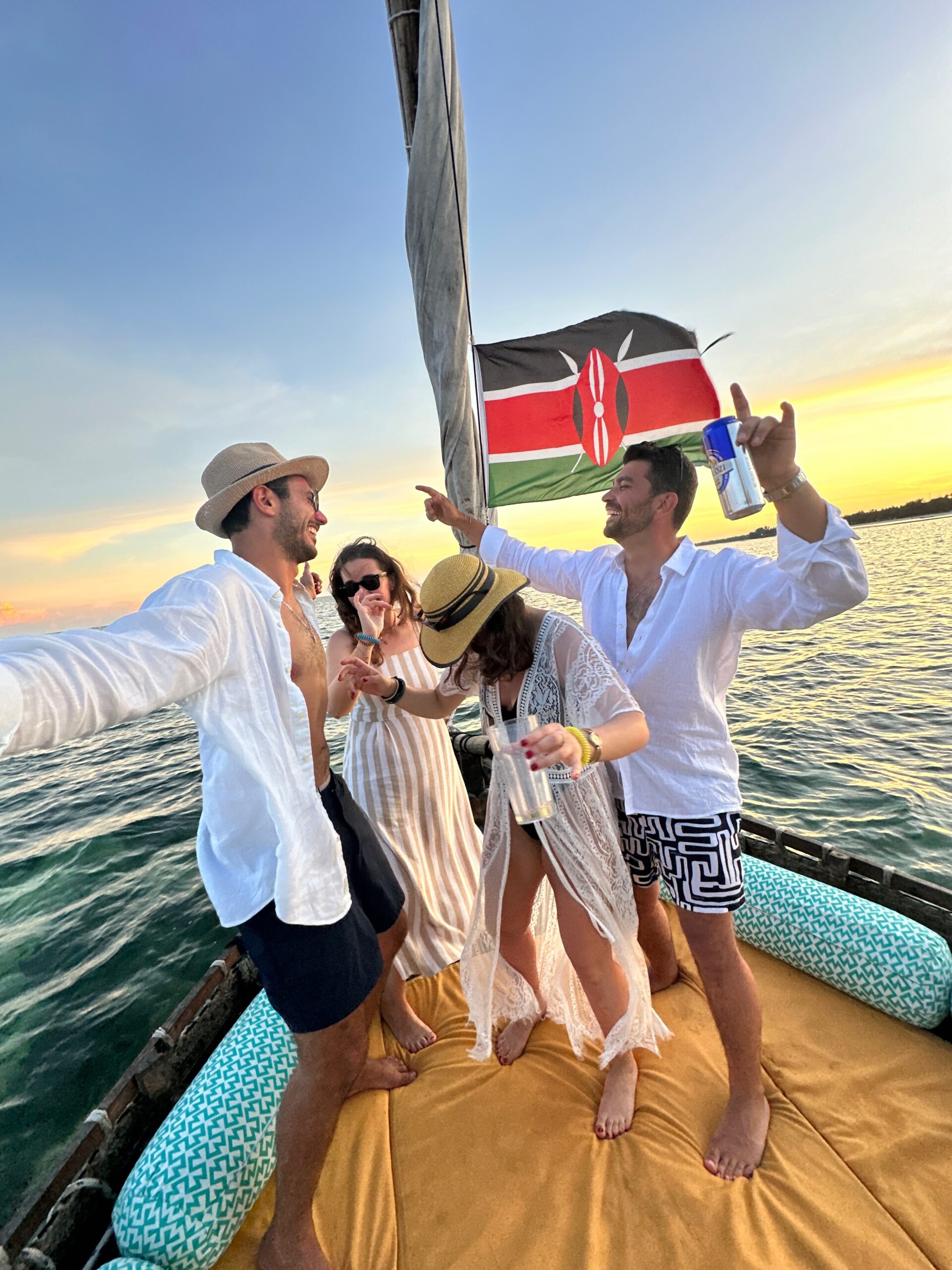 Culture Quest Kenya (7) A group of people enjoying a boat ride, dancing and holding drinks. They are under a Kenyan flag with a sunset over the water in the background.