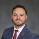 Headshot of a person wearing a blue suit, white shirt, and red tie, with a brooch on the lapel, against a neutral background.