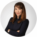 Headshot of person with long brown hair, wearing a dark blazer, arms crossed, and smiling with a neutral background.