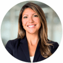 Headshot of person with long hair wearing a dark blazer, smiling and standing in a professional setting.