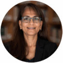 Headshot of a person wearing glasses and a dark blazer, smiling. Books are visible in the blurred background.