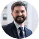 Headshot of person with a beard, wearing a suit and tie, smiling, in an urban setting.