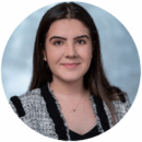 Headshot of person with long hair, wearing a patterned blazer and necklace, set against a soft-focus background.