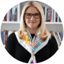 Headshot of a person with glasses, blonde hair, and a scarf, standing in front of a bookshelf.