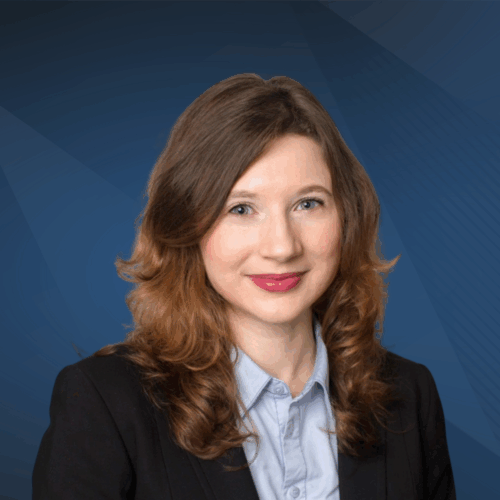 Sonia Alb_Jacobson Headshot of a person with long, wavy hair wearing a suit jacket and collared shirt, set against a dark blue background.